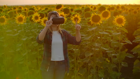 The-young-girl-with-long-hair-in-plaid-shirt-and-jeans-is-working-in-VR-glasses.-She-is-engaged-in-the-working-process.-It-is-a-perfect-sunny-day-in-the-sunflower-field.